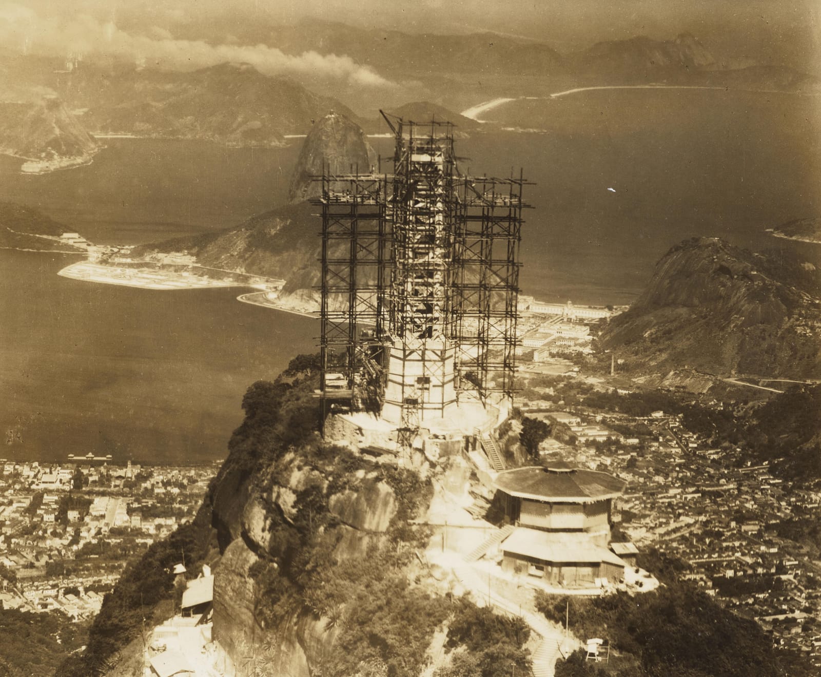 Christ the Redeemer under construction, wrapped in scaffolding on the peak of Corcovado, with Sugarloaf and the bay behind