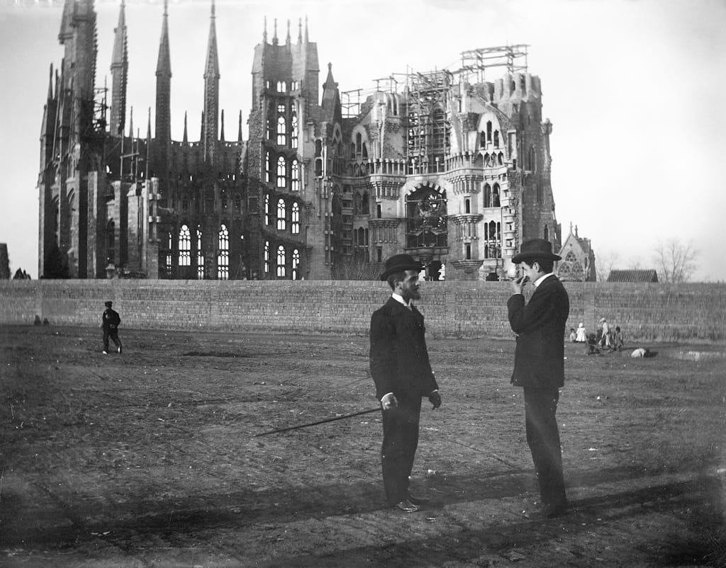 The Sagrada Família in 1905, half built under scaffolding behind a wall, with two men in bowler hats on open ground in front